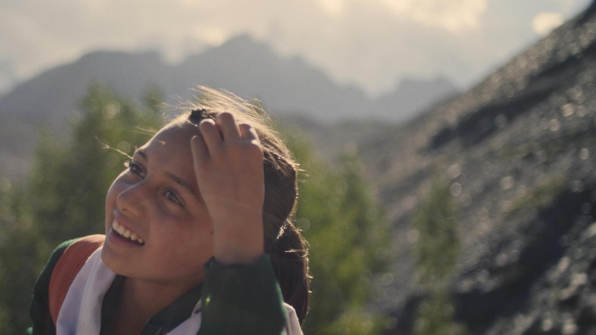 Une jeune fille sourit en se coiffant sur un fond montagneux ;  toujours du film 
